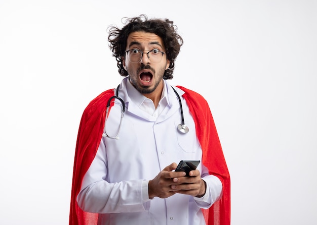 Anxious young caucasian superhero man in optical glasses wearing doctor uniform with red cloak and with stethoscope around neck holds phone and looks  with copy space