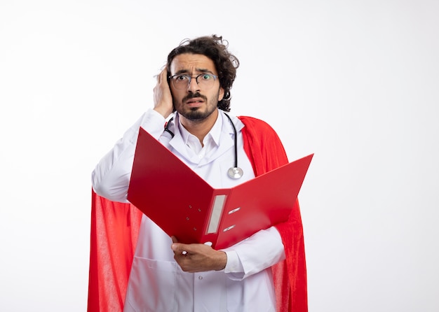 Anxious young caucasian superhero man in optical glasses wearing doctor uniform with red cloak and with stethoscope around neck puts hand on head and holds file folder isolated on white wall