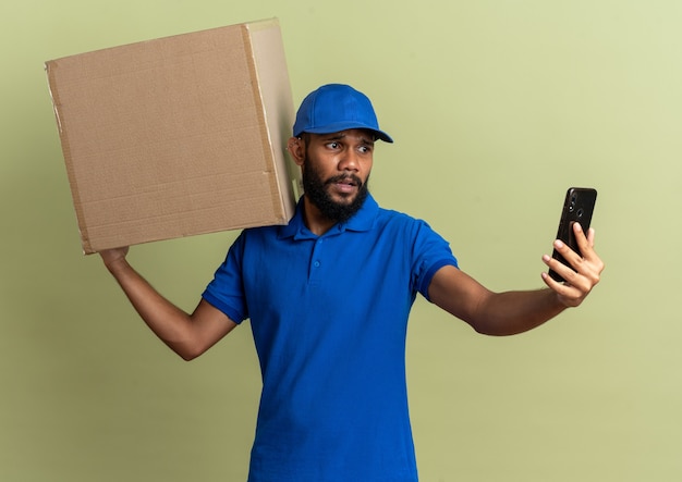 anxious young delivery man holding cardboard box and looking at phone isolated on olive green wall with copy space