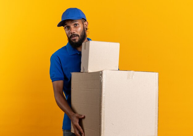 anxious young delivery man holding cardboard boxes isolated on orange wall with copy space