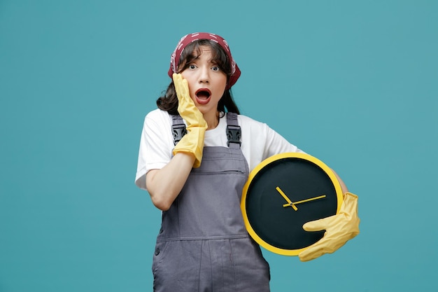 Foto gratuita ansiosa giovane donna addetta alle pulizie che indossa bandana uniforme e guanti di gomma che tengono l'orologio tenendo la mano sul viso guardando la fotocamera isolata su sfondo blu