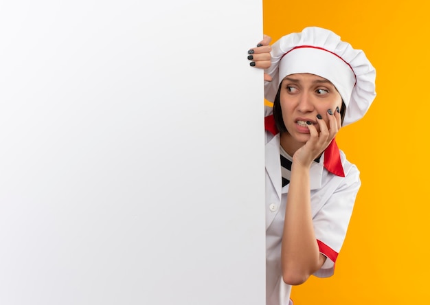 Anxious young female cook in chef uniform standing behind white wall and looking at side with hand on cheek isolated on orange  with copy space