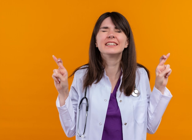 Anxious young female doctor in medical robe with stethoscope crosses fingers of both hands on isolated orange background with copy space