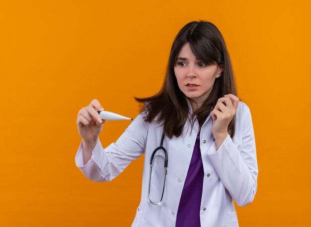 Anxious young female doctor in medical robe with stethoscope holds thermometer on isolated orange background with copy space