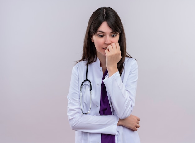 Anxious young female doctor in medical robe with stethoscope puts hand on chin and looks down on isolated white background with copy space
