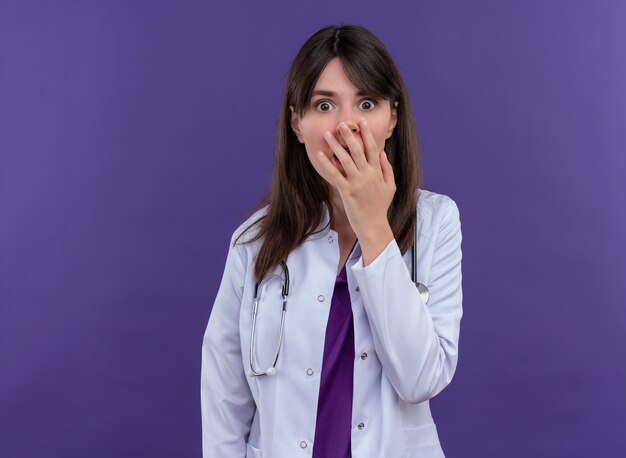 Anxious young female doctor in medical robe with stethoscope puts hand on mouth on isolated violet background with copy space