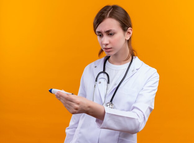 Anxious young female doctor wearing medical robe and stethoscope and holding thermometer looking at it on isolated orange space