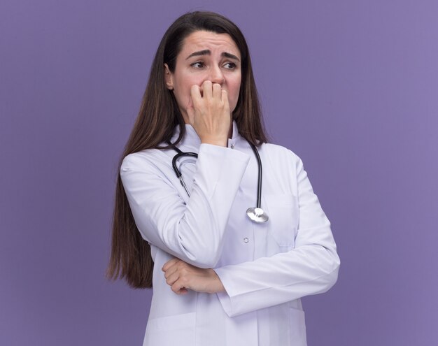 Тревожная молодая женщина-врач в медицинском халате со стетоскопом кусает ногти, глядя в сторону на фиолетовом