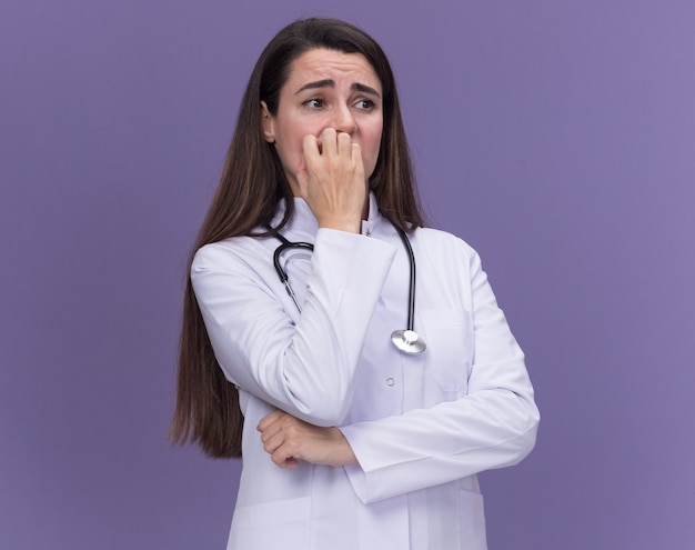 Anxious young female doctor wearing medical robe with stethoscope bites nails looking at side on purple 