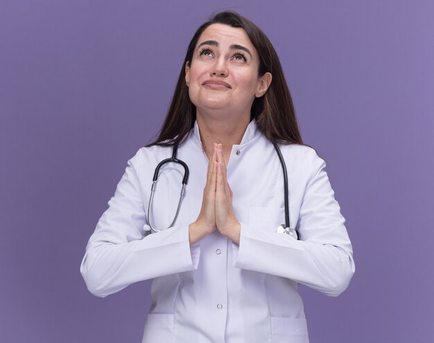 Тревожная молодая женщина-врач в медицинском халате со стетоскопом держит руки вместе и смотрит вверх на фиолетовый