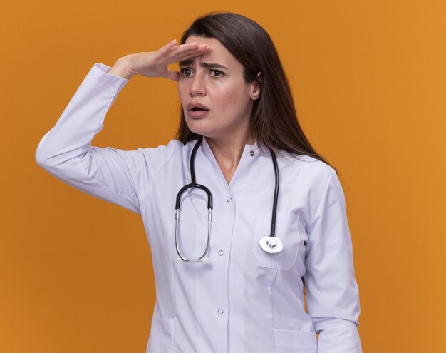 聴診器で医療ローブを身に着けている気になる若い女性医師は、オレンジ色の側面を見て額に手のひらを保ちます