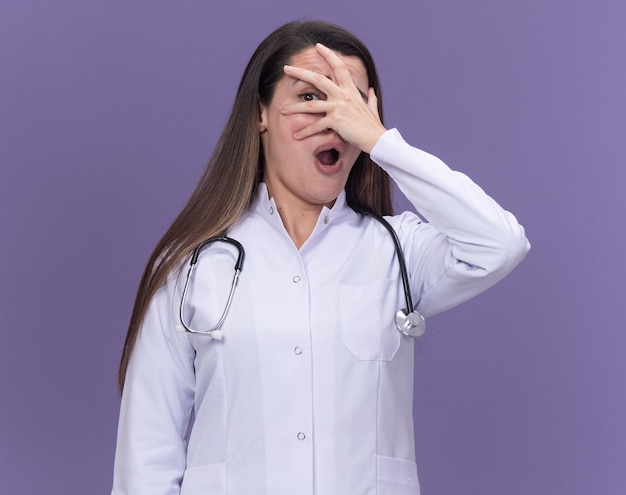 Anxious young female doctor wearing medical robe with stethoscope puts hand on face through fingers isolated on purple wall with copy space