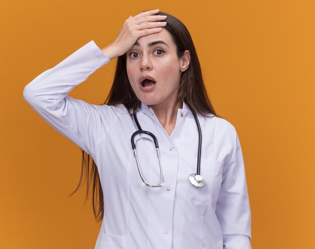 Anxious young female doctor wearing medical robe with stethoscope puts hand on forehead isolated on orange wall with copy space