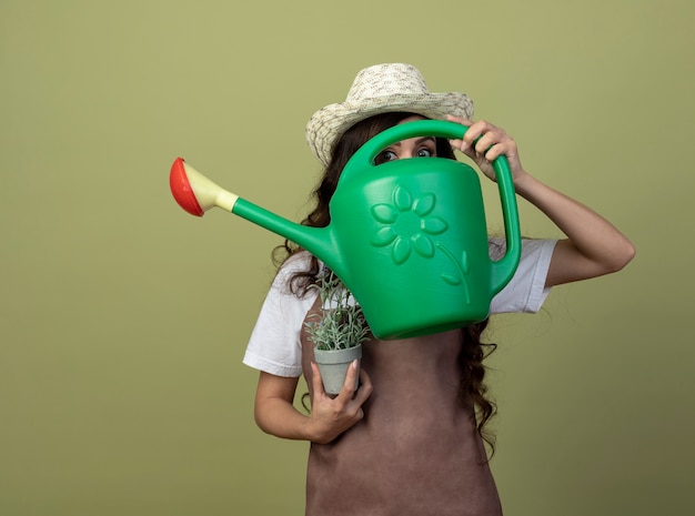 Anxious young female gardener in uniform wearing gardening hat holds flowerpot and looks at front through watering can isolated on olive green wall