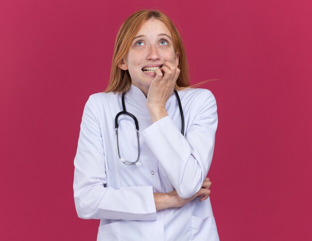 Anxious young female ginger doctor wearing medical robe and stethoscope putting hand on lip looking up isolated on crimson wall with copy space