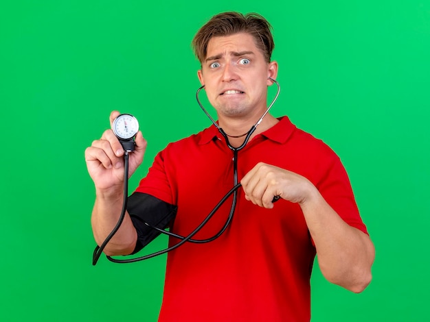 Anxious young handsome blonde ill man wearing stethoscope measuring pressure to himself holding sphygmomanometer looking at front isolated on green wall