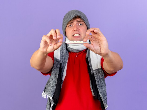 Anxious young handsome blonde ill man wearing winter hat and scarf stretching out thermometer towards camera looking at camera isolated on purple background