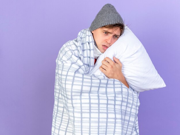 Anxious young handsome blonde ill man wearing winter hat wrapped in plaid holding pillow putting head on it looking straight isolated on purple background with copy space