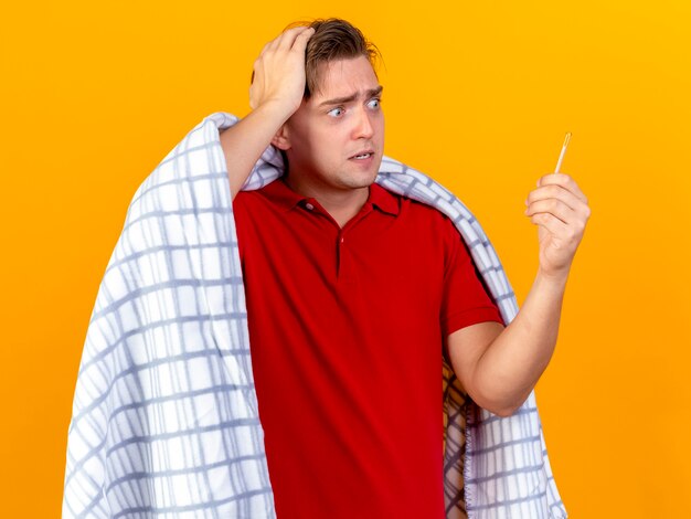 Anxious young handsome blonde ill man wrapped in plaid keeping hand on head holding and looking at thermometer isolated on orange wall