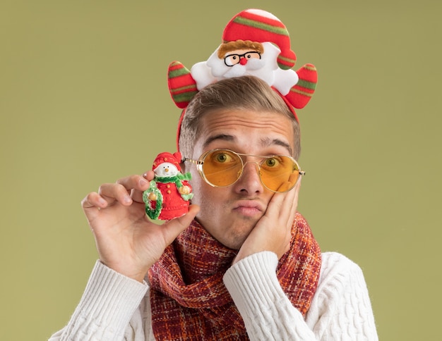 Foto gratuita ansioso giovane bel ragazzo che indossa la fascia di babbo natale e sciarpa guardando la telecamera tenendo pupazzo di neve ornamento di natale tenendo la mano sul viso isolato su sfondo verde oliva