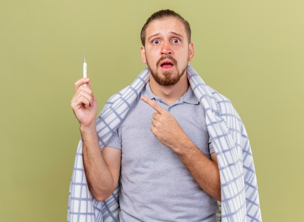 Anxious young handsome ill man wrapped in plaid holding and pointing at thermometer looking at front isolated on olive green wall