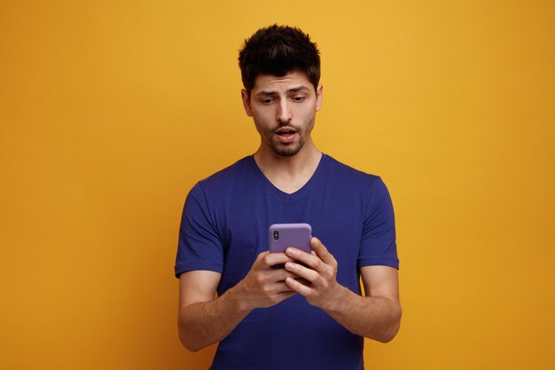 Anxious young handsome man holding and looking at his mobile phone on yellow background