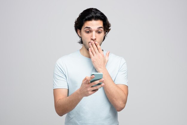 Тревожный молодой красивый мужчина держит и смотрит на мобильный телефон, держа руку на рту изолированы на белом фоне