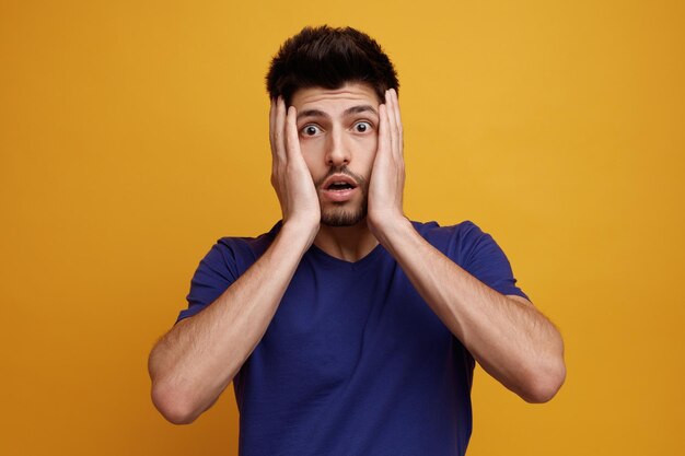 Anxious young handsome man looking at camera keeping hands on face on yellow background