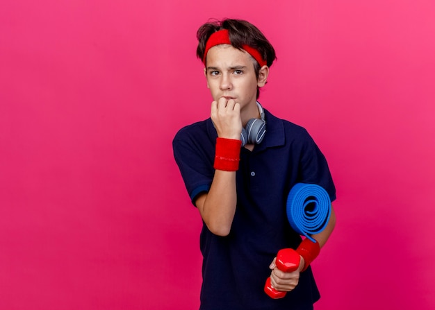 Free photo anxious young handsome sporty boy wearing headband and wristbands and headphones on neck, holding dumbbell with towel, touching chin isolated