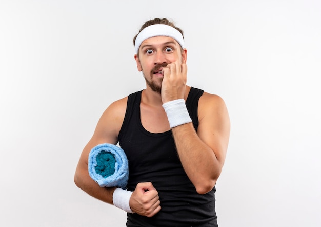 Anxious young handsome sporty man wearing headband and wristbands holding towel and biting his fingers isolated on white wall with copy space
