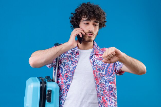 Anxious young handsome traveler man talking on phone with clenched fist looking at his hand with suitcase on isolated blue space