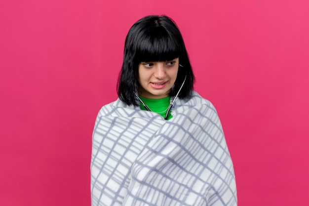 Anxious young ill caucasian girl wrapped in plaid wearing stethoscope looking at side isolated on crimson wall with copy space