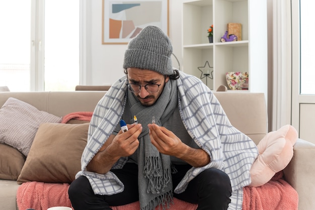 Free photo anxious young ill caucasian man in optical glasses wrapped in plaid with scarf around his neck wearing winter hat holding thermometer syringe and medical ampoule sitting on couch at living room