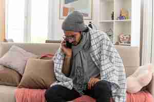 Бесплатное фото Встревоженный молодой больной человек в оптических очках, завернутый в плед с шарфом на шее, в зимней шапке разговаривает по телефону, сидя на диване в гостиной
