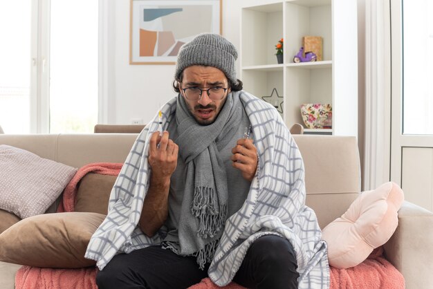 anxious young ill man in optical glasses wrapped in plaid with scarf around his neck wearing winter hat holding medical ampoule and looking at syringe sitting on couch at living room