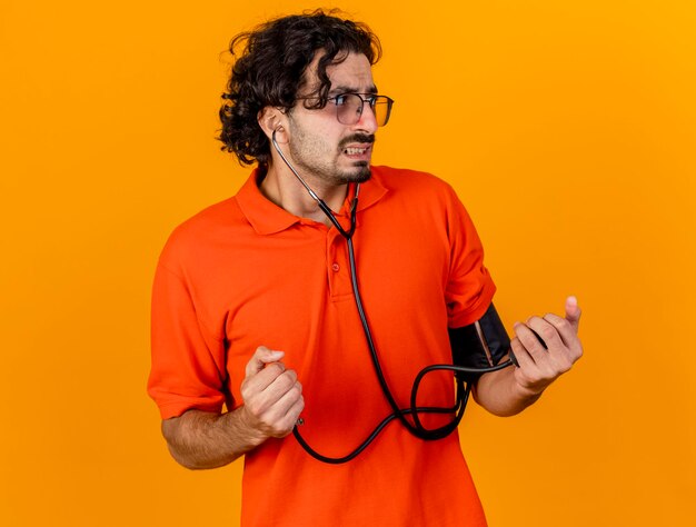 Anxious young ill man wearing glasses and stethoscope measuring his pressure with sphygmomanometer looking at side isolated on orange wall