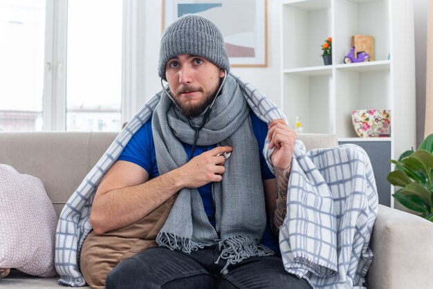 Anxious young ill man wearing scarf and winter hat with stethoscope wrapped in blanket sitting on sofa in living room looking up listening to his own heartbeats