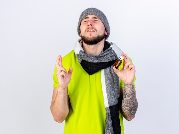 Anxious young ill man wearing winter hat and scarf crosses fingers and holds medicine in a glass bottle isolated on white wall