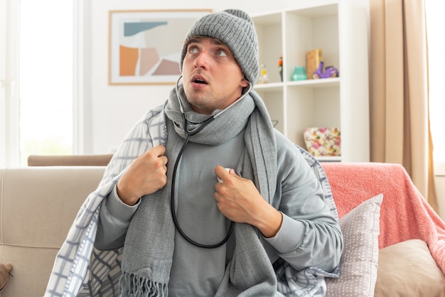 anxious young ill man with scarf around neck wearing winter hat wrapped in plaid measuring his heart beat with stethoscope looking at side sitting on couch at living room