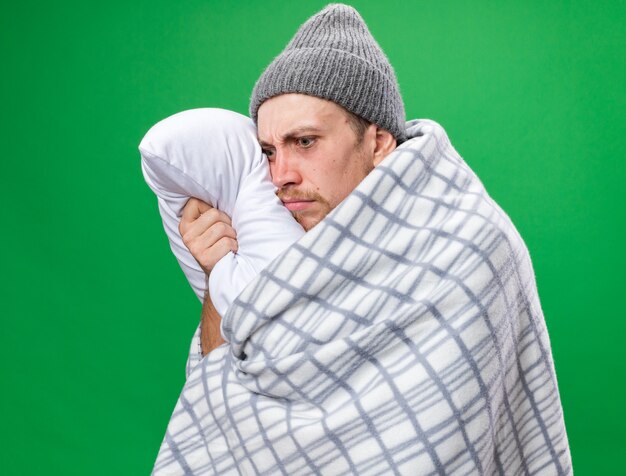 Anxious young ill slavic man with scarf around neck wrapped in plaid wearing winter hat holds pillow isolated on green wall with copy space