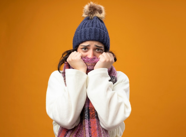Бесплатное фото Тревожная молодая больная женщина в зимней шапке и шарфе смотрит на передний прикрывающий рот шарфом, изолированным на оранжевой стене