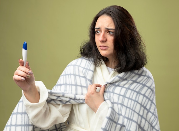 Anxious young ill woman wearing robe wrapped in plaid holding and looking at thermometer grabbing plaid isolated on olive green wall