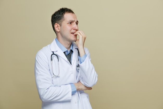 anxious young male doctor wearing medical robe and stethoscope around neck keeping hand under elbow looking at side while biting fingers isolated on olive green background with copy space