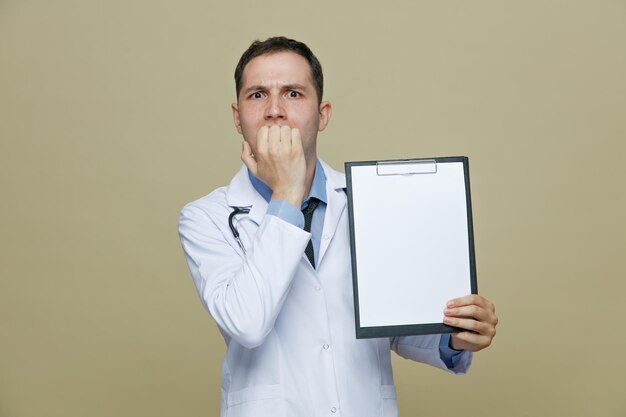 Тревожный молодой врач-мужчина в медицинском халате и стетоскопе на шее показывает буфер обмена, глядя на камеру, кусая пальцы на оливково-зеленом фоне