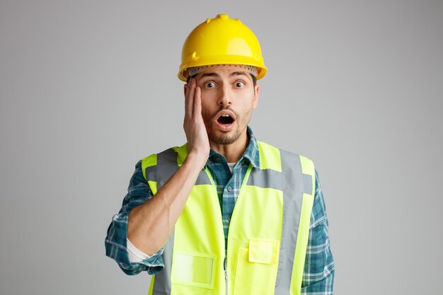 Anxious young male engineer wearing safety helmet and uniform keeping hand on face looking at camera isolated on white background