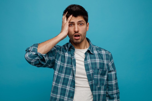 Anxious young man keeping hand on head looking at camera isolated on blue background