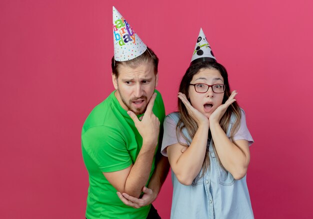 Anxious young man wearing party hat puts hand on chin looking down and standing with shocked young girl wearing party hat holding hands on chin isolated on pink wall