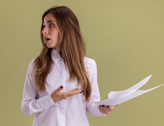 Anxious young pretty caucasian girl holds and points at blank paper sheets looking at side on olive green 