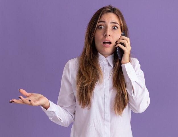 Anxious young pretty caucasian girl keeps hand open and talks on phone on purple 