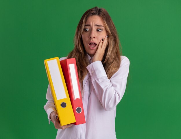 Anxious young pretty caucasian girl puts hand on face holding and looking at file folders on green 